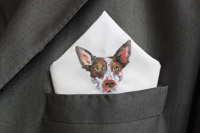 Close-up of the hand embroidery: a brown and white dog face with amber eyes and pink-lined ears stitched in thread on a white pocket square.