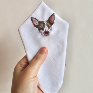 Hand holding a white cotton handkerchief with a small embroidered dog portrait on the folded corner, showing the size of the design.