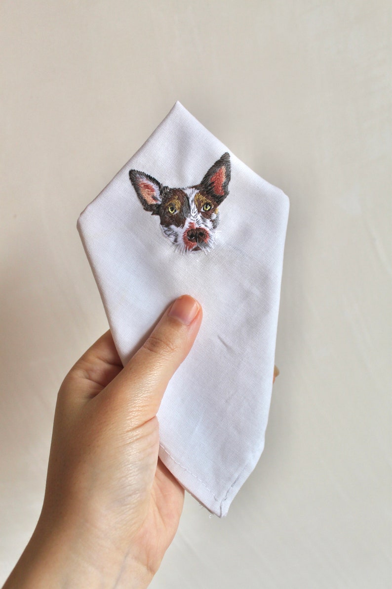 Hand holding a white cotton handkerchief with a small embroidered dog portrait on the folded corner, showing the size of the design.