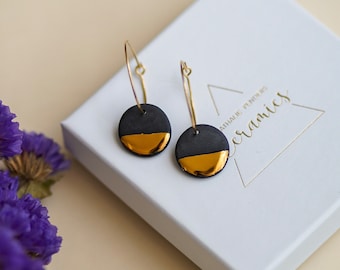 May include: A pair of black and gold hoop earrings with a gold accent on the bottom half of the circle. The earrings are on a white background with a white box that says "ATHALIE TYMONS CERAMICS".