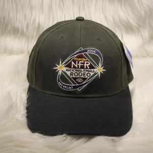 Op de afbeelding: Een donker olijfgroene honkbalpet met een zwarte klep. De voorkant is voorzien van een geborduurd logo voor de 2004 Wrangler National Finals Rodeo in Las Vegas, afgebeeld met een ruitvorm, steraccenten en blauwe gebogen tekstelementen.
