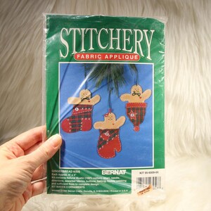 以下が含まれることがあります： BernatのStitcheryファブリックアップリケキット「Gingerbread Kids」。透明なプラスチック袋に入っています。緑色のパッケージには、赤と緑のチェック柄の靴下とミトンに入った3つのジンジャーブレッドのオーナメントが描かれています。