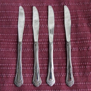 May include: Five silver butter knives with ornate handles featuring a rose design. The knives are arranged in a row on a red and white patterned tablecloth.