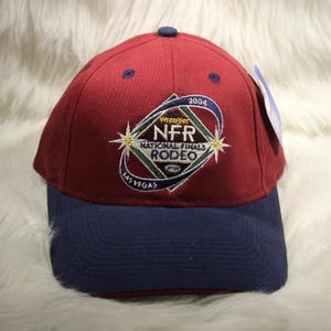 Op de afbeelding: Een bordeauxrode honkbalpet met een marineblauwe klep en een knoop aan de bovenkant. De voorkant is voorzien van een geborduurd logo voor de 2004 Wrangler National Finals Rodeo in Las Vegas, inclusief een ruitvorm, steraccenten en de tekst NFR National Finals Rodeo.