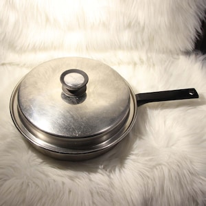 May include: A stainless steel skillet with a lid and a black handle. The lid has a small, round handle. The skillet is round and appears to be made of a durable metal. The handle is attached to the side of the skillet.