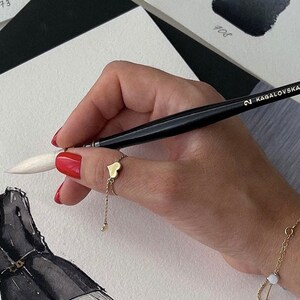 May include: A gold heart ring with a chain and a gold bracelet with a small white bead are visible. The hand holds a black paintbrush with the text "KAGALOVSKA by daVinci". The image also shows a fashion illustration with a woman wearing sunglasses.