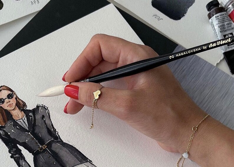 May include: A gold heart ring with a chain and a gold bracelet with a small white bead are visible. The hand holds a black paintbrush with the text "KAGALOVSKA by daVinci". The image also shows a fashion illustration with a woman wearing sunglasses.