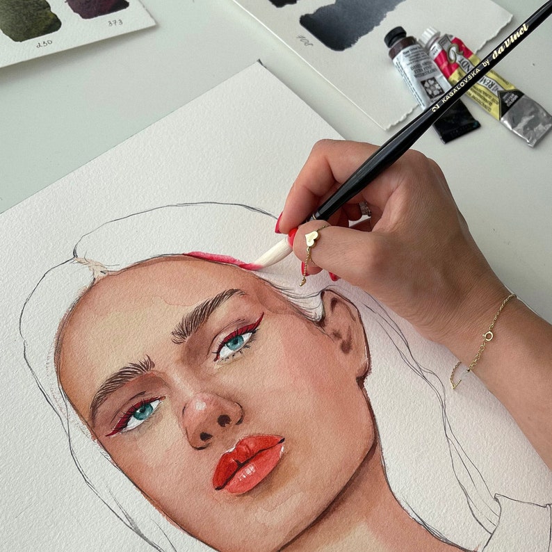 May include: A watercolor painting of a woman's face with red eyeliner and lipstick. A hand holding a paintbrush is adding red paint to the woman's hair. Watercolor paints and a paintbrush are on the white surface.