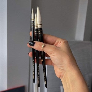 May include: Three black watercolor paint brushes with white bristles and gold accents are held in a hand. The brushes are labeled "KAGALOVSKA by da Vinci" with sizes 0, 2, and 4. The brushes are for painting.