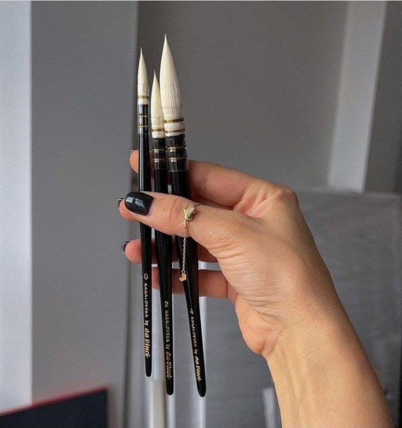 May include: Three black watercolor paint brushes with white bristles and gold accents are held in a hand. The brushes are labeled "KAGALOVSKA by da Vinci" with sizes 0, 2, and 4. The brushes are for painting.