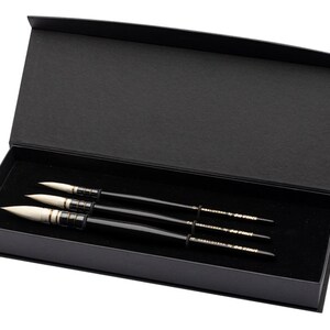 May include: A set of three black handled paint brushes with white tips, presented in a black box with a hinged lid. The brushes are arranged neatly on a black velvet surface.