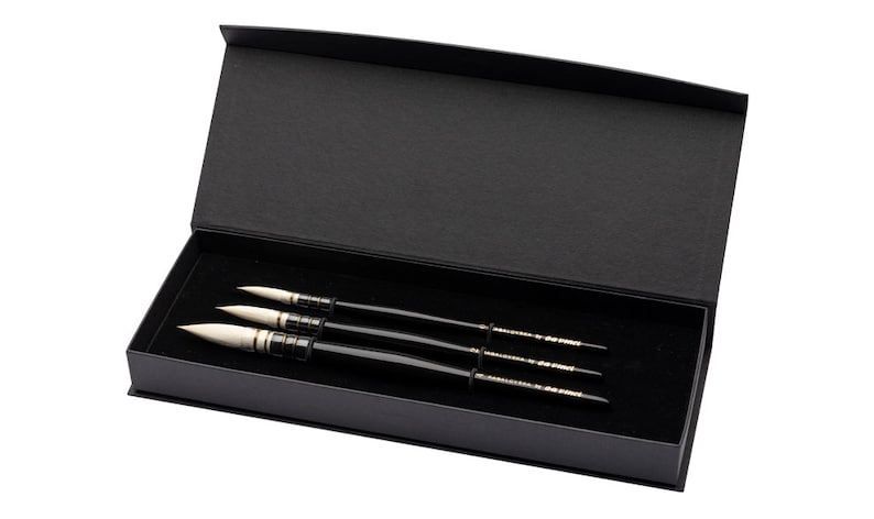 May include: A set of three black handled paint brushes with white tips, presented in a black box with a hinged lid. The brushes are arranged neatly on a black velvet surface.