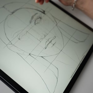 May include: A close-up of a digital drawing of a face on a tablet screen. The face is drawn with a grid overlay, and the artist is using a stylus to draw the details.