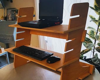 Puede incluir: Un escritorio de pie de madera de dos niveles. Un portátil negro está en el nivel superior, con un teclado y un ratón negros en el nivel inferior. El escritorio es de madera clara y está sobre una mesa redonda.