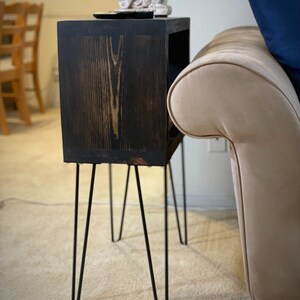 Könnte beinhalten: Ein schwarzer Holz-Beistelltisch mit einem kleinen Regal. Der Tisch hat vier Haarnadelbeine und steht auf einem beigefarbenen Teppich. Eine kleine weiße Figur steht auf dem Tisch.