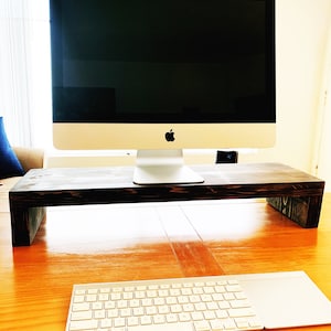 May include: A dark wood monitor stand with a silver iMac computer on top. A white wireless keyboard and trackpad are on the wooden desk below the stand.