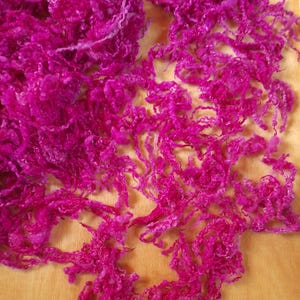 Op de afbeelding: Close-up van levendig fuchsia-kleurig garen, met een getextureerde, krullende uitstraling. Het garen is gerangschikt in losse klonten, wat de zachte, donzige aard laat zien. De achtergrond is een licht houten oppervlak.