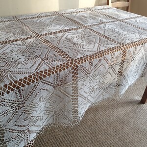 May include: White crocheted tablecloth with a delicate pattern. The tablecloth is laid on a wooden table and has a fringed edge.