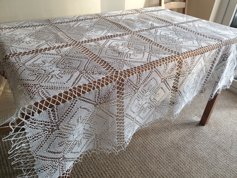 May include: White crocheted tablecloth with a delicate pattern. The tablecloth is laid on a wooden table and has a fringed edge.