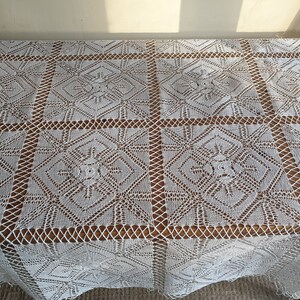 May include: White crocheted tablecloth with a delicate floral pattern. The tablecloth is laid out on a wooden table.
