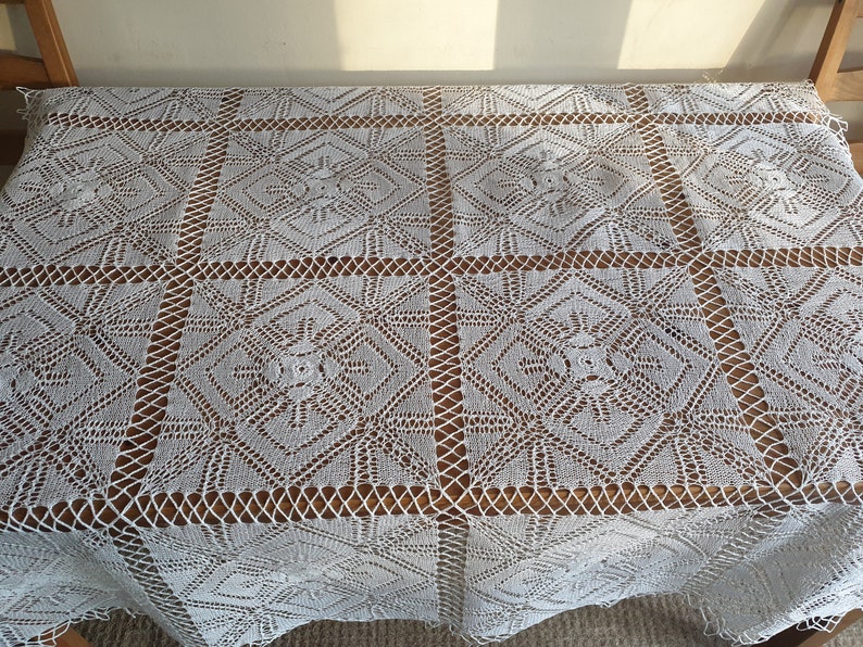 May include: White crocheted tablecloth with a delicate floral pattern. The tablecloth is laid out on a wooden table.