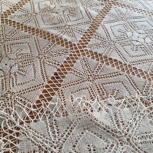 May include: White crocheted tablecloth with a geometric pattern. The tablecloth has a delicate, lacy design with a square shape.