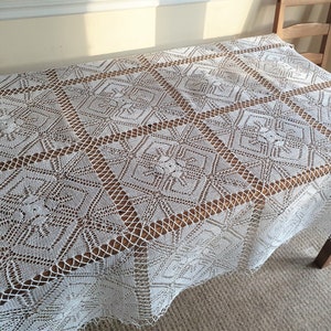 May include: White crocheted tablecloth with a square pattern. The tablecloth is laid on a wooden table.