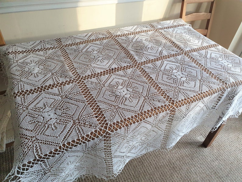 May include: White crocheted tablecloth with a square pattern. The tablecloth is laid on a wooden table.