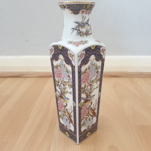 Può includere: Un vaso in porcellana bianca con un motivo floreale blu e oro. Il vaso ha una base quadrata e un collo stretto.