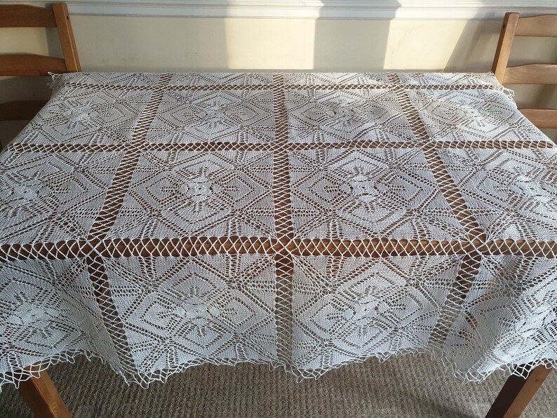 May include: White crocheted tablecloth with a geometric pattern. The tablecloth is laid out on a wooden table.