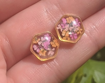 May include: Two hexagonal stud earrings with gold-coloured frames. Each earring features small, dried flowers in pink, orange, and white, set in clear resin. The earrings are shown on a person's hand, with green foliage in the background.