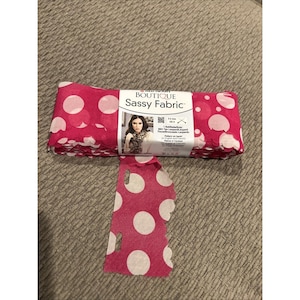 Könnte beinhalten: Eine Rolle Red Heart Boutique Sassy Fabric Garn in leuchtendem Pink mit großen weißen Punkten. Der Stoff hat einen gewellten Rand und vorgestanzte Löcher zum Stricken. Das Etikett zeigt den Text Red Heart Boutique Sassy Fabric.