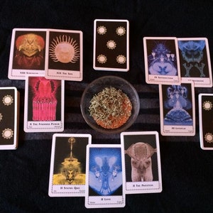 May include: A tarot card reading layout featuring several cards with intricate illustrations and text. The cards are arranged around a small bowl containing a dried plant material. The cards have a black background with white borders and symbols.