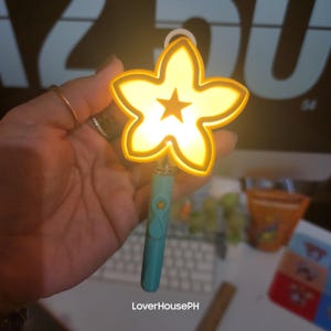May include: A star-shaped, yellow light-up wand with a small star in the center. The wand has a teal handle with a decorative design. The image includes the text "LoverHousePH".