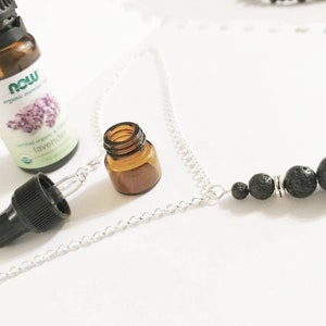 Puede incluir: Un collar de cadena plateada con cuentas de piedra de lava negra de varios tamaños. También hay una pequeña botella de vidrio ámbar y una botella de aceite esencial de lavanda, junto con un cuentagotas.