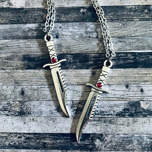 May include: Two silver knife pendant necklaces with red gem accents. The necklaces are on a wood background.