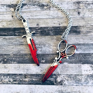 May include: Two silver necklaces with red accents. One necklace features a knife pendant and the other features a pair of scissors pendant.