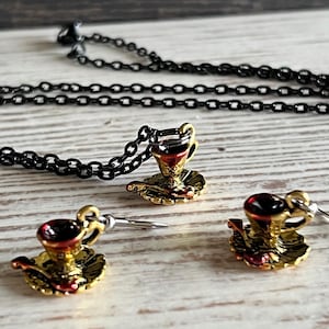 May include: A set of gold and red teacup and saucer jewelry. The set includes a necklace with a teacup pendant and a pair of earrings with teacup charms.