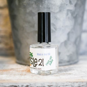 May include: A clear glass bottle of peel-off base coat nail polish with a black cap. The bottle has a white label with green palm trees and the text "Isle 21" and "Peel Off Base" on it. The label also says "Made in the USA" and "www.isle21.com".