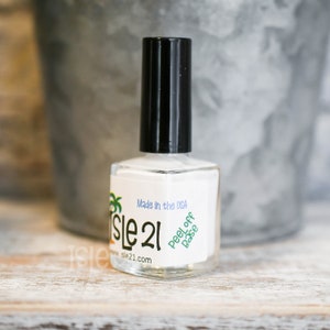May include: A clear glass bottle of white nail polish with a black cap. The label reads "Isle 21" and "Peel Off Base" with a palm tree graphic. The bottle is made in the USA.