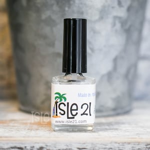 May include: A clear glass bottle with a black cap and a white label. The label has a green palm tree and the text "Isle 21" and "www.isle21.com". The bottle is sitting on a white wooden surface.