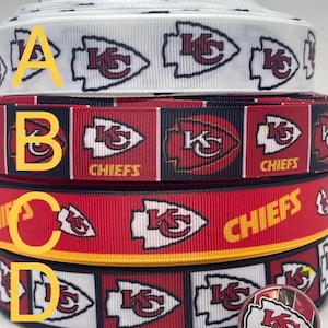 Puede incluir: Varios rollos de cinta blanca, negra y roja con un patrón repetido de un logotipo de equipo. También se ve un botón con el mismo logotipo. El texto en la parte superior dice "Available in 7/8" & 1.5".