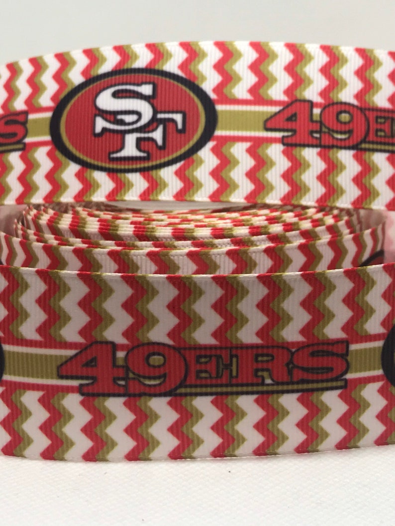 以下が含まれることがあります： サンフランシスコ・フォーティナイナーズのロゴと「49ers」の文字が入った、赤、白、金のヘリンボーン柄のリボンです。
