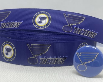 Pode incluir: Um rolo de fita azul marinho e um botão a condizer com o logótipo dos St. Louis Blues. A fita tem logótipos e texto repetidos em branco e dourado. O botão é redondo e tem um acabamento brilhante.