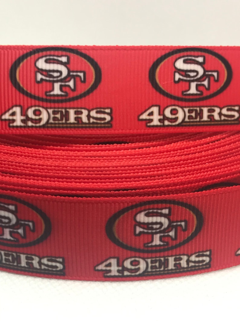 以下が含まれることがあります： サンフランシスコ・フォーティナイナーズのロゴと「49ers」という文字が繰り返された赤と金のリボンです。