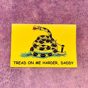 Puede incluir: Un rectángulo amarillo con una ilustración de una serpiente enrollada. La serpiente es negra y amarilla, con una lengua roja. El texto "TREAD ON ME HARDER, DADDY" está impreso en negro en la parte inferior del rectángulo.