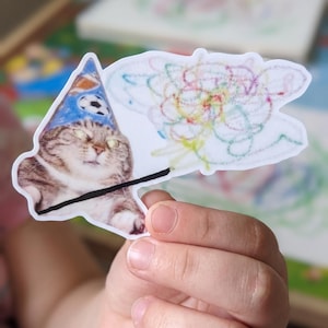 May include: A die-cut sticker featuring a brown tabby cat wearing a blue party hat with a soccer ball pattern. The cat is holding a black wand that extends into a colorful, multi-colored scribble drawing of a flower-like shape on a white background.