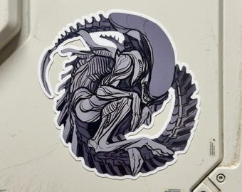 May include: A circular sticker featuring a detailed illustration of a Xenomorph creature in shades of grey and purple. The creature is curled into a fetal position, showcasing its elongated head, segmented tail, and ribbed exoskeleton structure.