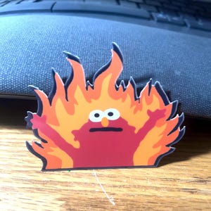 May include: A sticker featuring a cartoon character engulfed in flames. The character is red with white eyes and a black mouth. The flames are orange and yellow, creating a fiery effect. The sticker is on a wooden surface.
