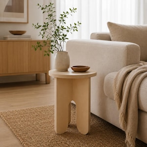 May include: A light-colored wooden side table with a circular top and a base featuring two arched legs. The table holds a speckled beige ceramic vase containing green leafy branches and a small, round, dark brown wooden bowl on its surface.
